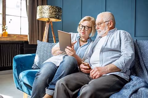 An elderly couple sitting closely together on a blue couch in a cozy living room, looking at a tablet device. The room features a large window with curtains, a decorative lamp with a patterned shade, and a blue paneled wall in the background.