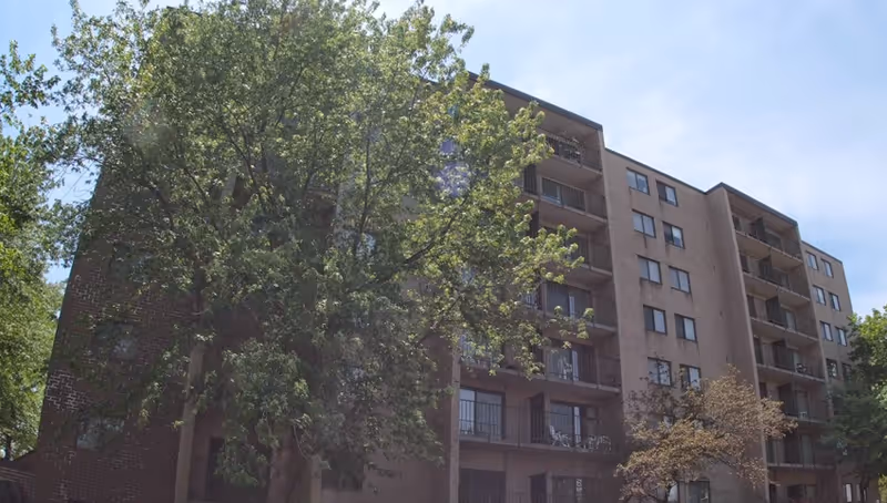 Exterior view of a multi-story senior living facility building partially obscured by leafy green trees under a clear blue sky.