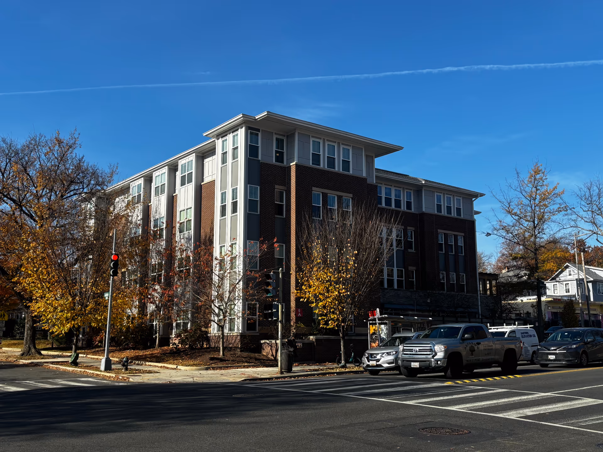 Exterior view of a multi-story senior living facility building named Maple Heights Senior Living, with a brick and white panel facade, surrounded by trees with autumn foliage, and several vehicles parked along the street in front.