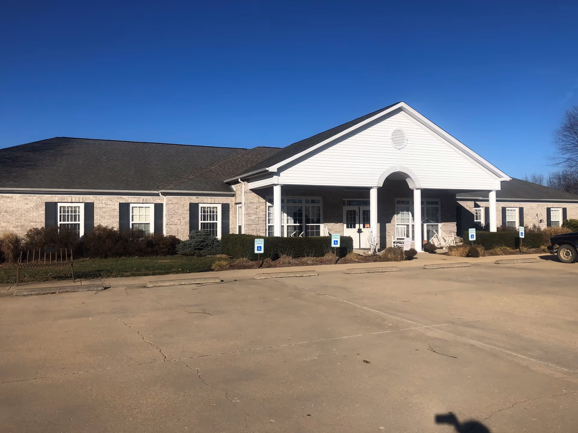 Exterior view of a single-story brick building with a white entrance porch supported by columns. There are several windows with black shutters and a parking area with handicapped parking signs in front. The sky is clear and blue.