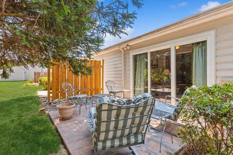 Outdoor patio area with metal chairs and a cushioned bench on a tiled floor next to a building with sliding glass doors and green curtains. There is a wooden fence, potted plant, green grass, and trees surrounding the patio under a blue sky with some clouds.