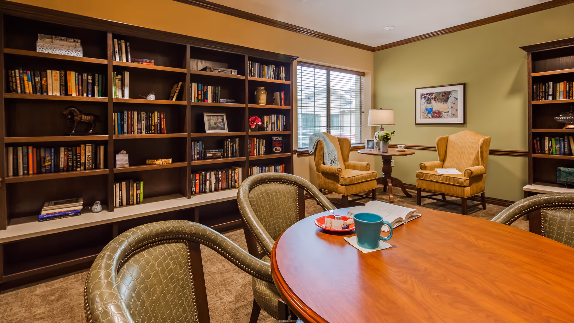 A cozy reading room with a large wooden bookshelf filled with books along the wall. In the foreground, there is a round wooden table with four green cushioned chairs. On the table, there is a blue mug, an open book, and a small plate with snacks. In the background, two yellow armchairs are positioned near a window with blinds, next to a small round table with a lamp and a framed picture on the wall.