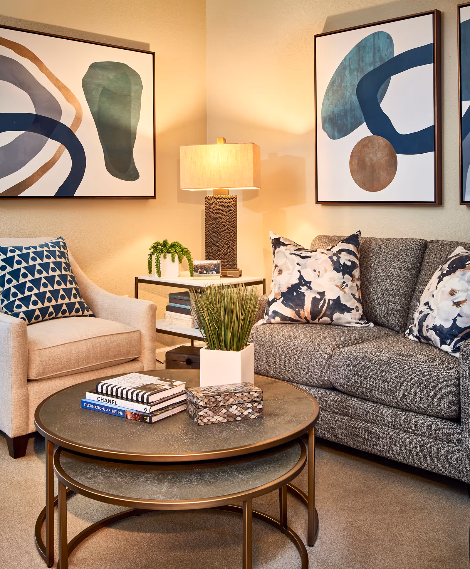 A cozy living room area featuring a gray upholstered sofa with floral patterned pillows, a beige armchair with a geometric patterned pillow, two nested round coffee tables with books and a decorative box, a small potted plant, and a side table with a lamp and another small plant. Abstract art pieces hang on the beige wall behind the furniture.