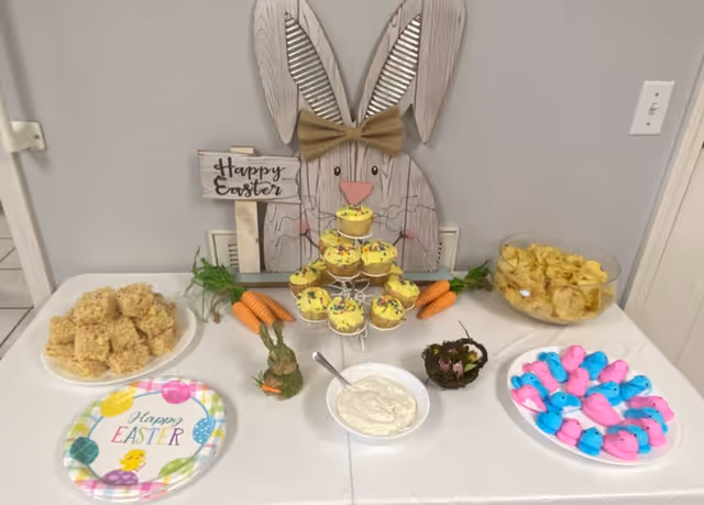 A festive Easter-themed snack table with a wooden bunny decoration wearing a bow tie and holding a 'Happy Easter' sign. The table has yellow frosted cupcakes on a tiered stand, a bowl of potato chips, a plate of pink and blue marshmallow peeps, a plate of rice crispy treats, a bowl of dip with a spoon, and decorative carrots and a small bunny figurine. A colorful 'Happy Easter' paper plate is also on the table.