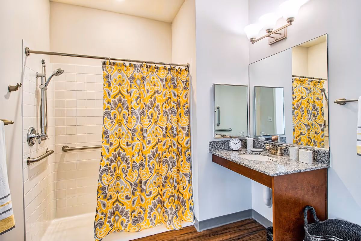 A clean and modern bathroom featuring a walk-in shower with a yellow and gray patterned shower curtain, grab bars for accessibility, a granite countertop with a sink, a large mirror, a small clock, and a basket on the floor.