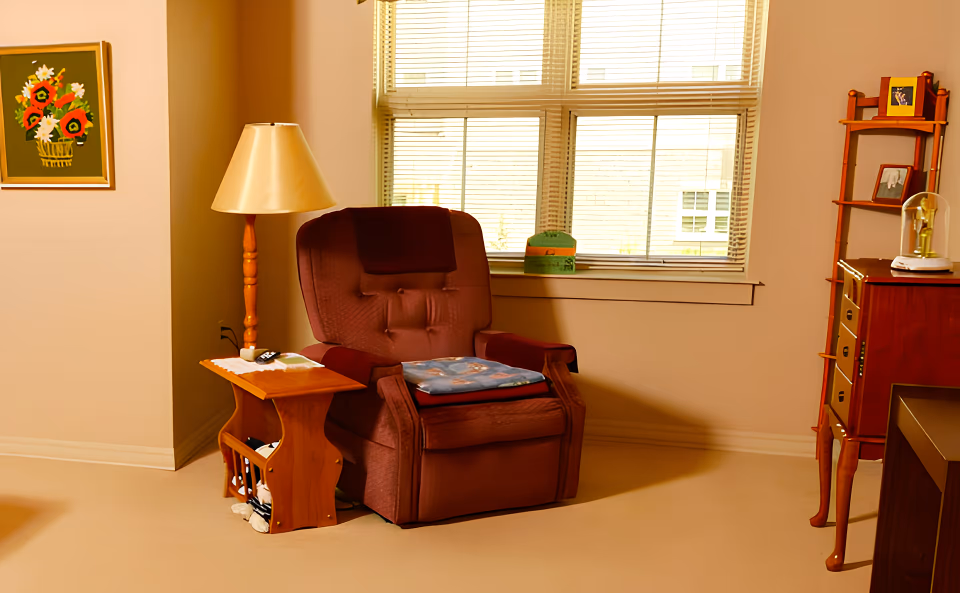 A cozy living room corner with a maroon recliner chair, a wooden side table holding a remote control and a lamp with a beige shade. Behind the chair is a window with blinds partially open, and to the right is a wooden shelving unit with framed photos and a decorative item under a glass dome. A framed painting of flowers hangs on the wall to the left.