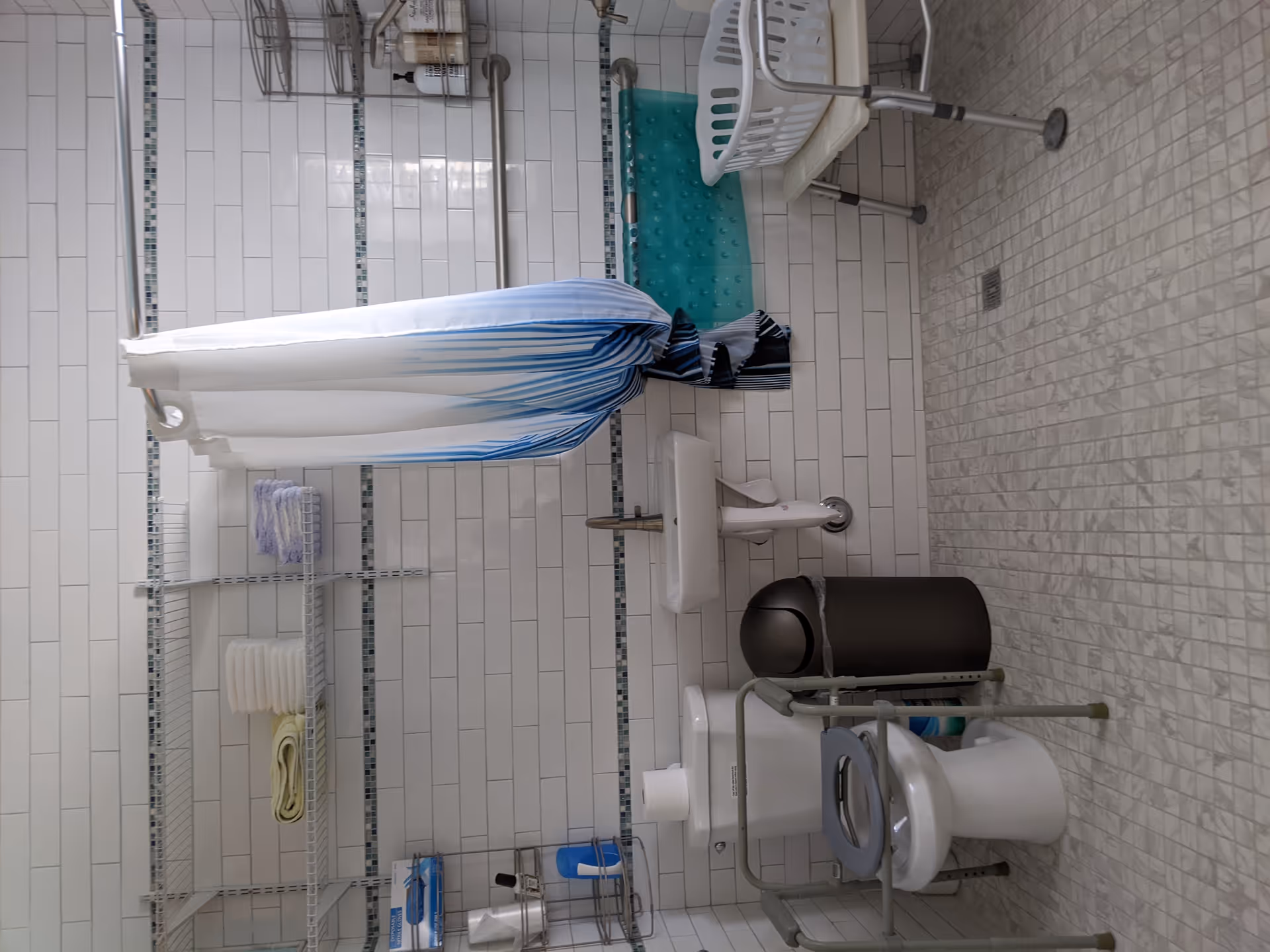 Accessible tiled bathroom with a shower and curtain, grab bars, a shower chair, raised toilet with support frame, sink, and shelving stocked with towels.