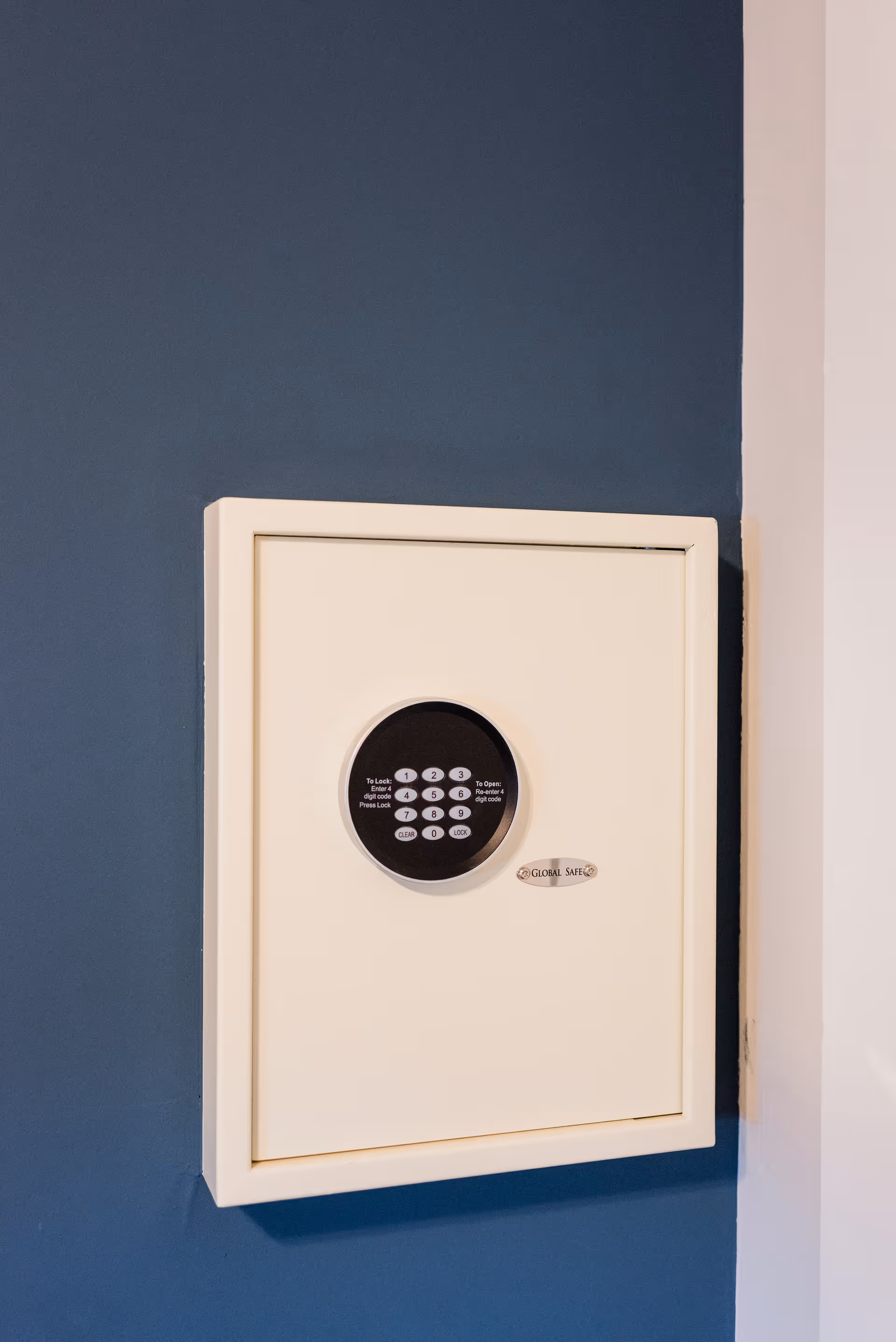 A beige wall-mounted safe with a black circular electronic keypad on a blue wall.
