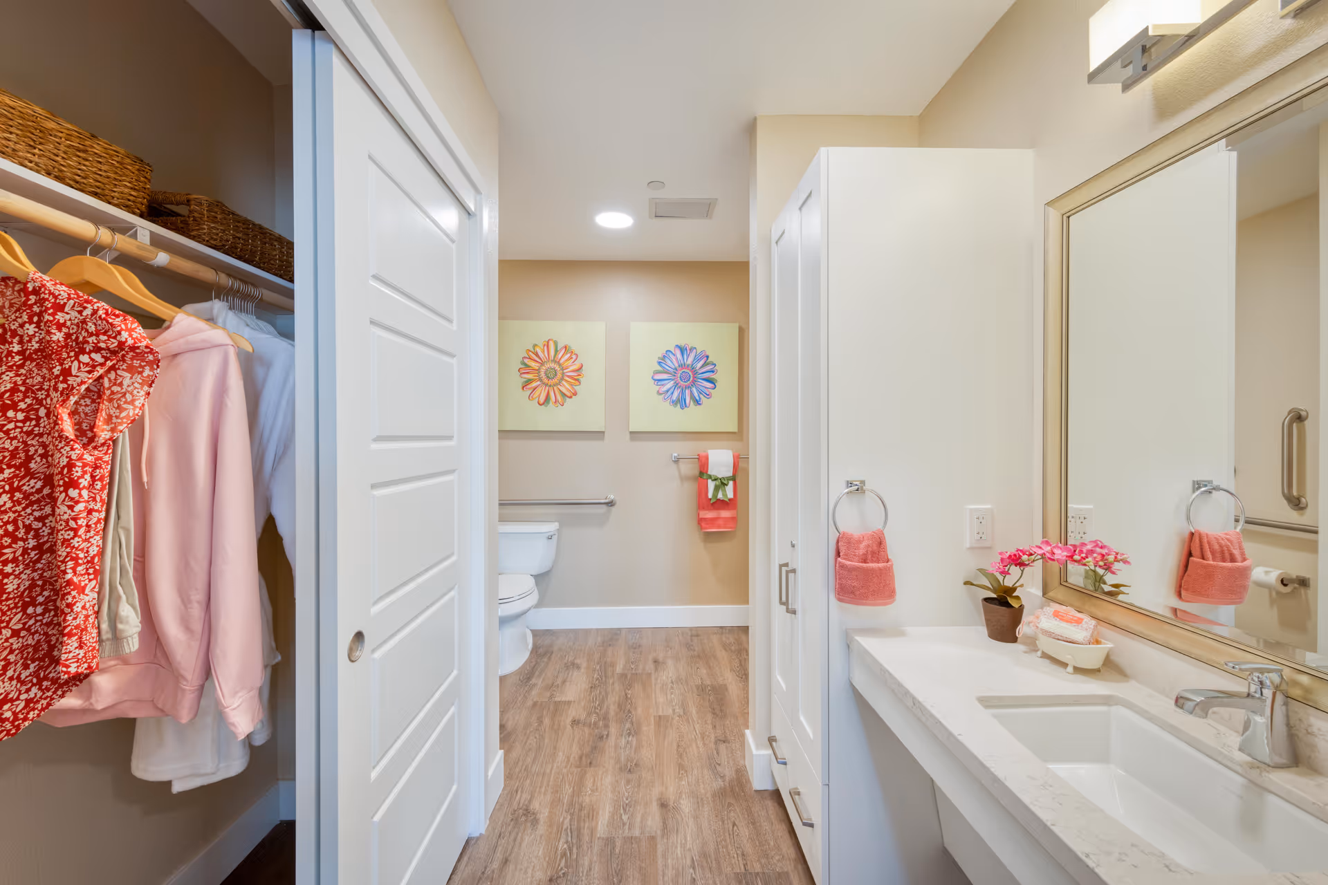 Bright, modern bathroom featuring a sink and mirror on the right, a toilet and floral wall art straight ahead, and an open closet with hanging clothes on the left.