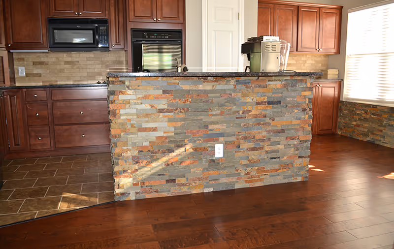 Interior view of a kitchen area with wooden cabinets, a built-in microwave and oven, and a kitchen island with a stone facade. The floor is a combination of tile and wood, and there is a window with blinds allowing natural light into the room.