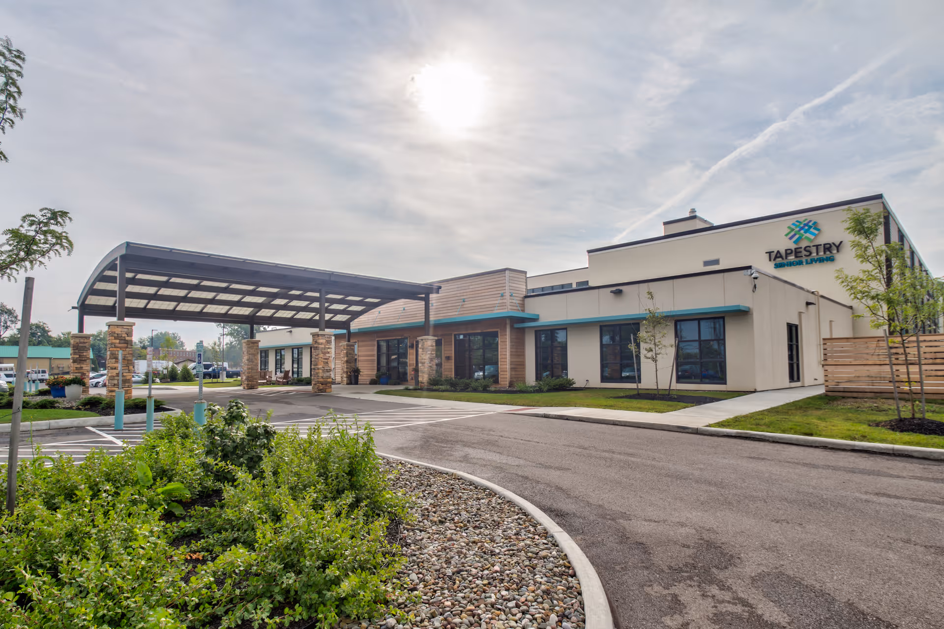 Exterior view of a modern senior living facility building with a covered entrance supported by stone pillars, landscaped greenery, and a clear sky with the sun partially covered by clouds. The building has the sign 'Tapestry Senior Living' on the upper right side.
