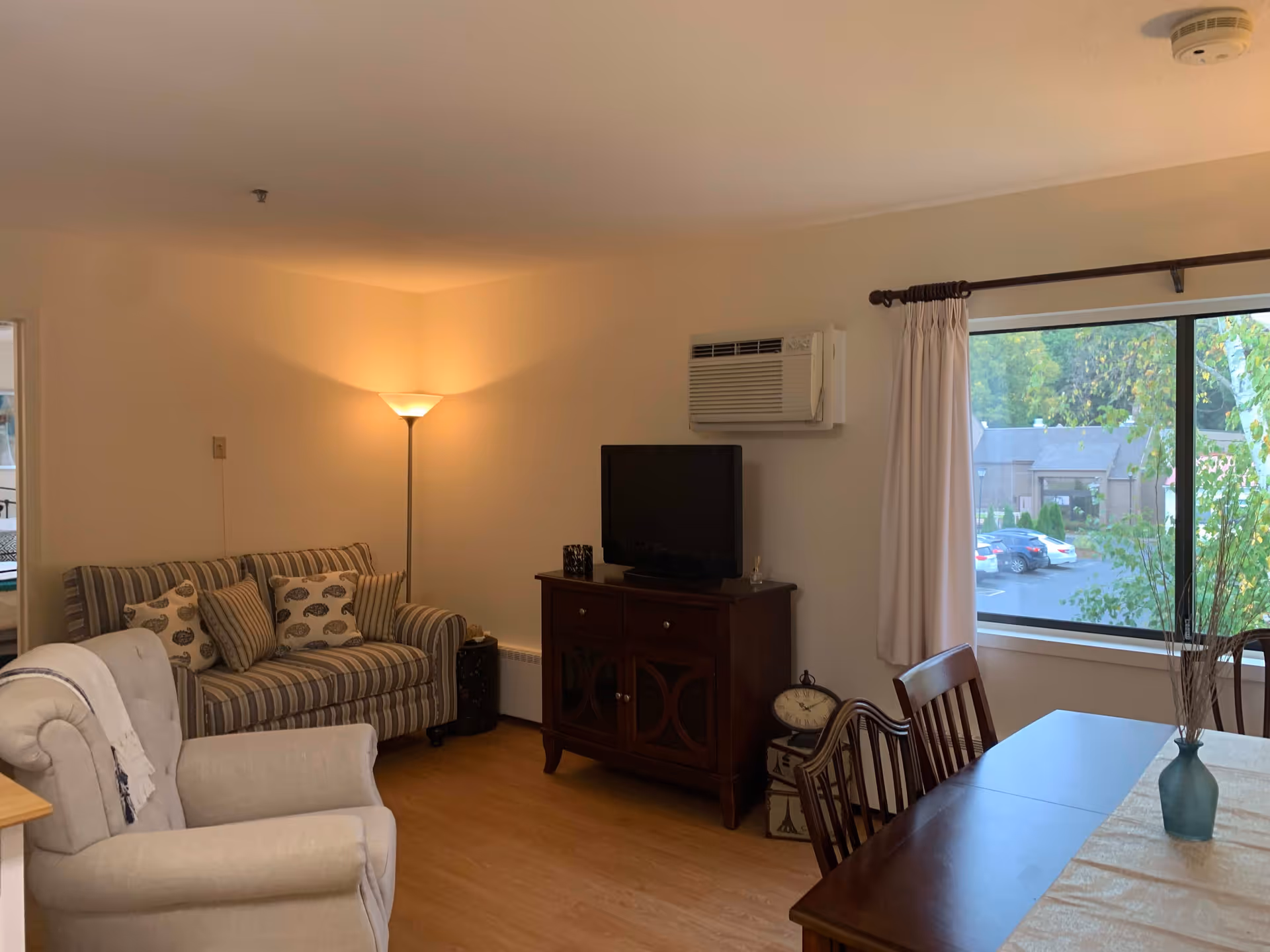 A cozy living room in a senior living facility with a striped loveseat, a cushioned armchair, a wooden TV stand with a flat-screen TV, a floor lamp in the corner, and a large window with white curtains overlooking a parking lot and trees. A wooden dining table with chairs and a vase with decorative branches is partially visible.