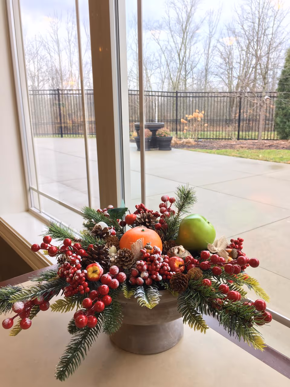 A festive centerpiece on a table near a large window, featuring pine branches, red berries, pine cones, an orange, a green apple, and other decorative elements. Outside the window, there is a paved patio area and a black metal fence with leafless trees in the background.