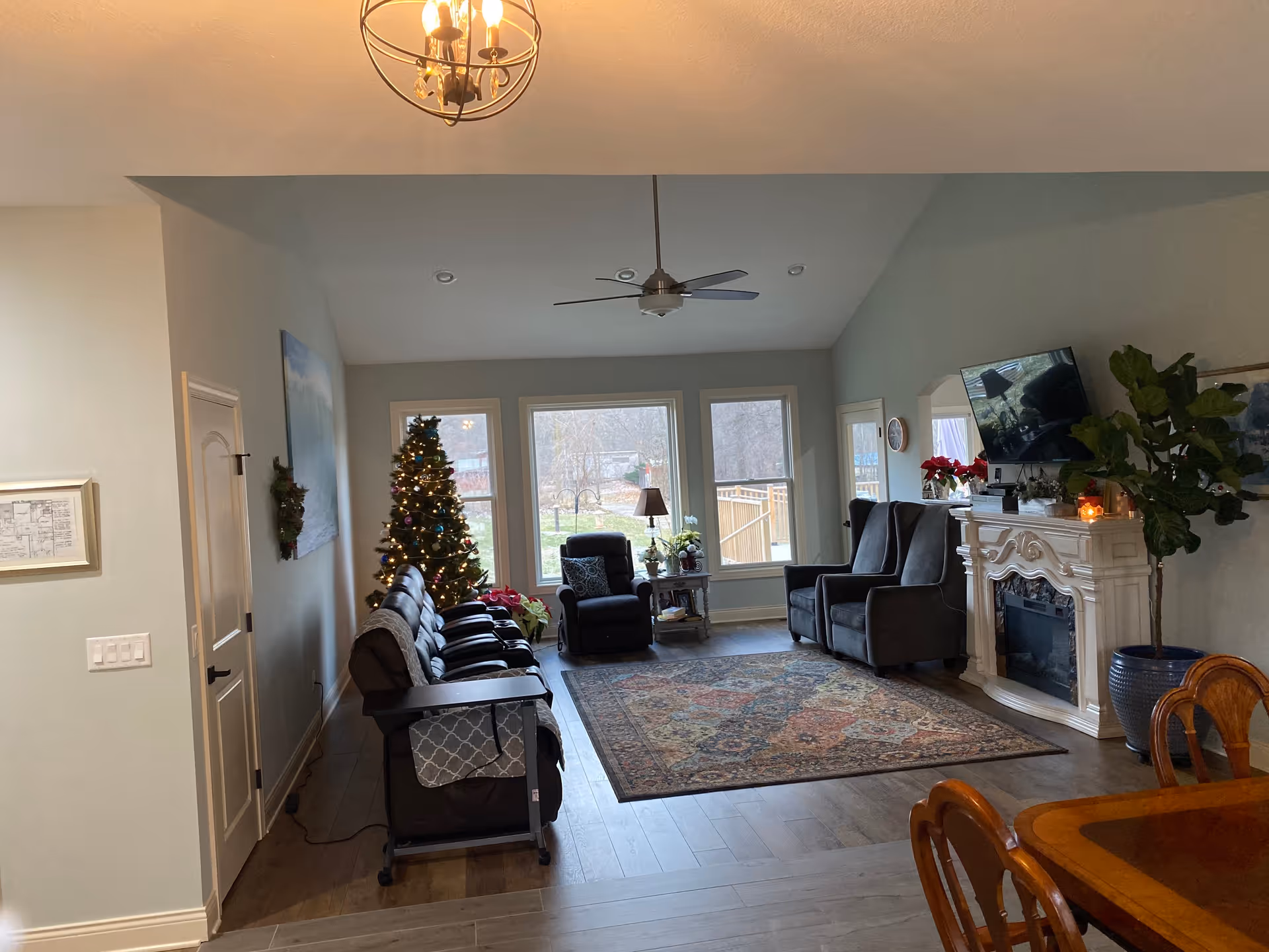 A cozy living room in Waltz Manor Senior Home featuring a decorated Christmas tree near large windows, several comfortable recliner chairs arranged around a patterned area rug, a white ornate fireplace with a mounted TV above it, and a wooden dining table partially visible in the foreground. The room has light-colored walls, a ceiling fan, and a chandelier.