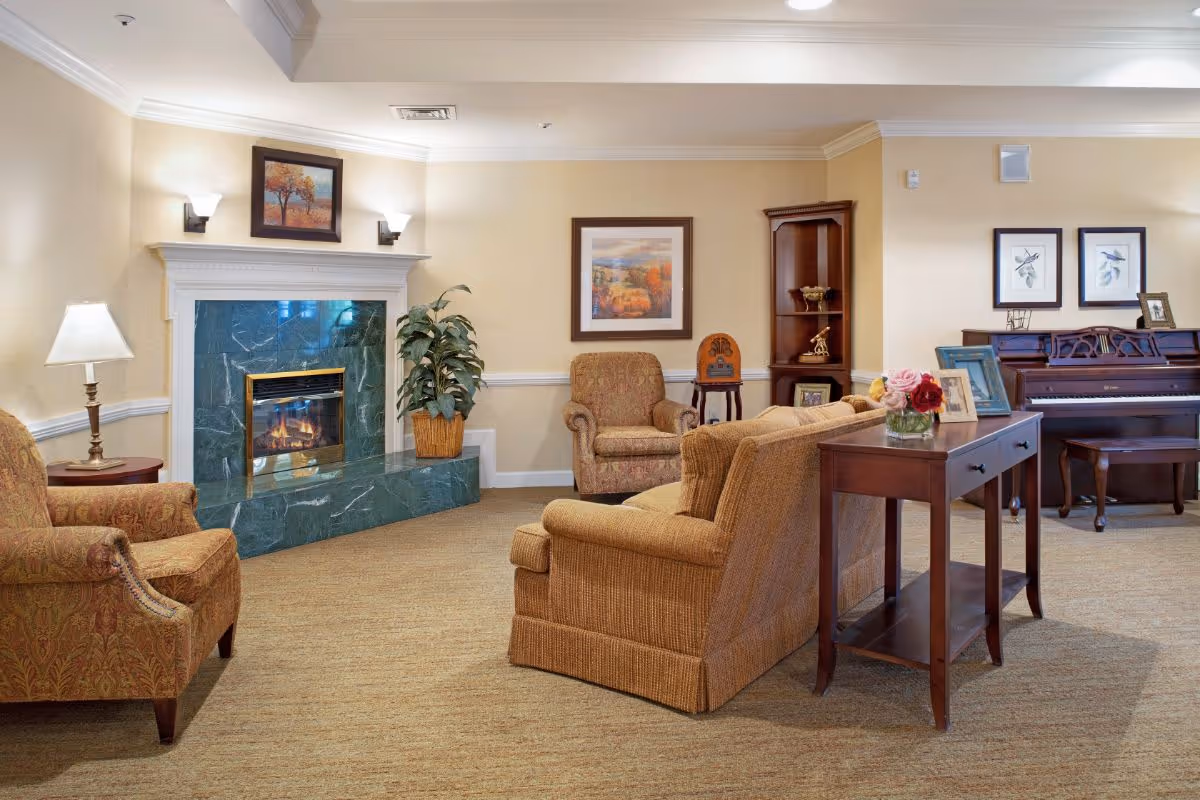 A cozy living room area with a green marble fireplace, two armchairs, a sofa, a wooden side table with a vase of flowers, a floor lamp, framed artwork on the walls, a potted plant, a wooden corner shelf with decorative items, and a piano with a bench.