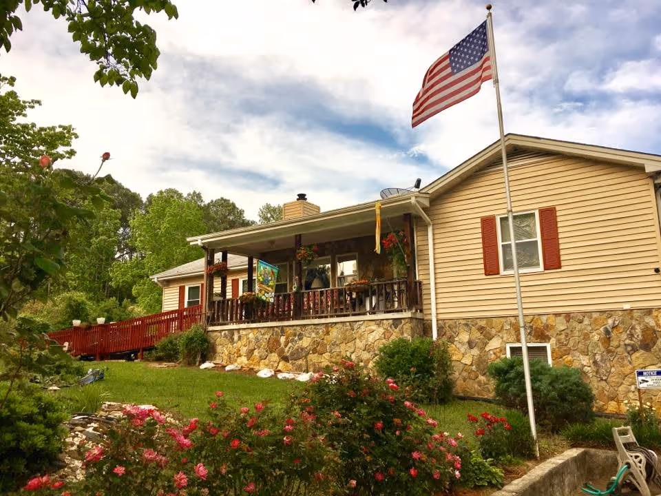 A single-story building with beige siding and a stone foundation, featuring a porch with hanging flower baskets and an American flag on a flagpole in the front yard. The yard has green grass, bushes, and blooming pink flowers under a partly cloudy sky.