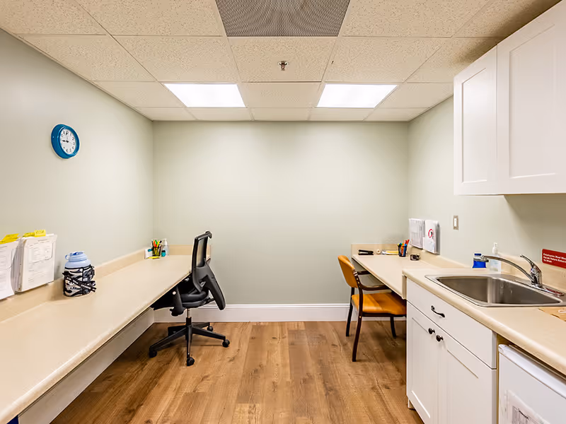A small office or workroom with light green walls, wood flooring, and a drop ceiling with fluorescent lights. There are two desks with chairs, office supplies, and paperwork. On the right side, there is a countertop with a sink and white cabinets above and below. A blue clock is mounted on the left wall.