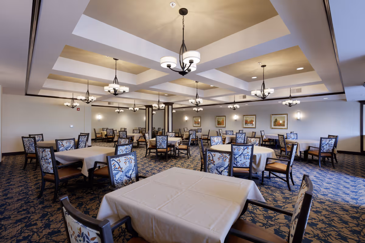 A spacious dining room with multiple tables covered in beige tablecloths and chairs with patterned upholstery. The ceiling features recessed lighting and several hanging chandeliers. The carpet has a blue and beige floral pattern, and framed artwork is displayed on the walls.