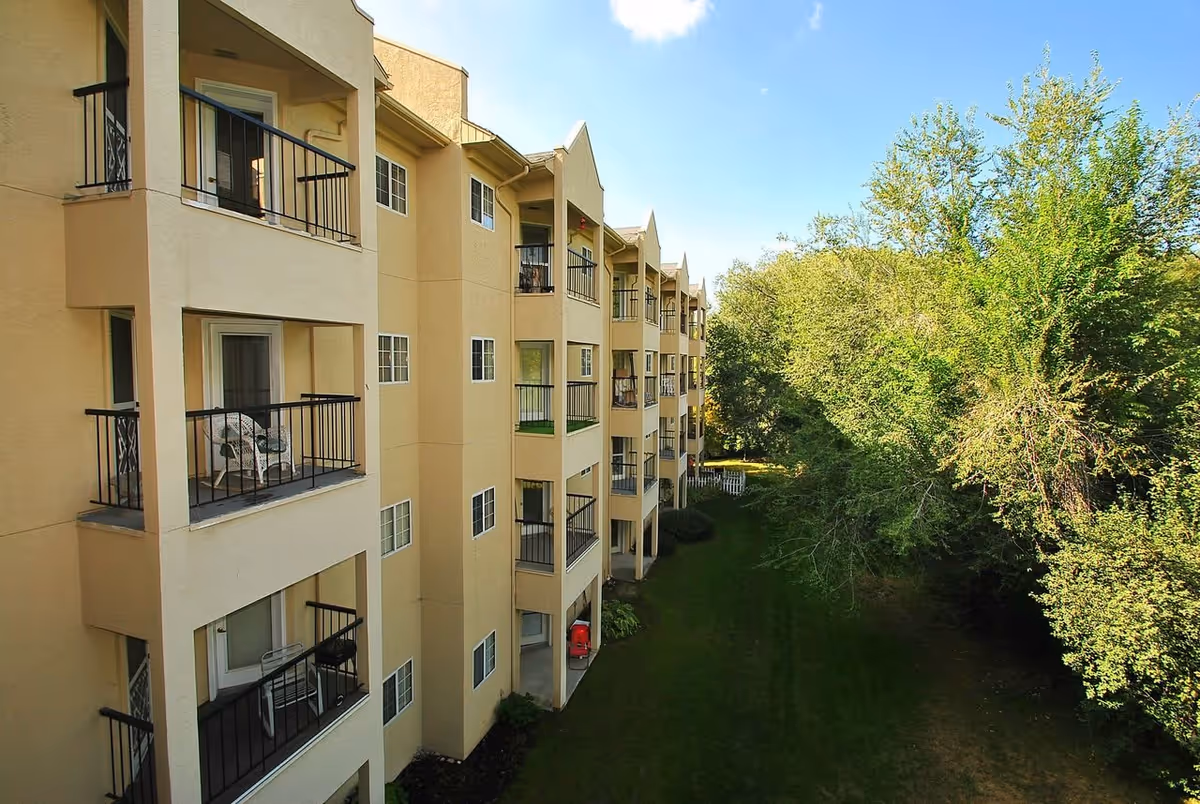Exterior view of a multi-story beige residential building with balconies overlooking a grassy area and trees under a clear blue sky.