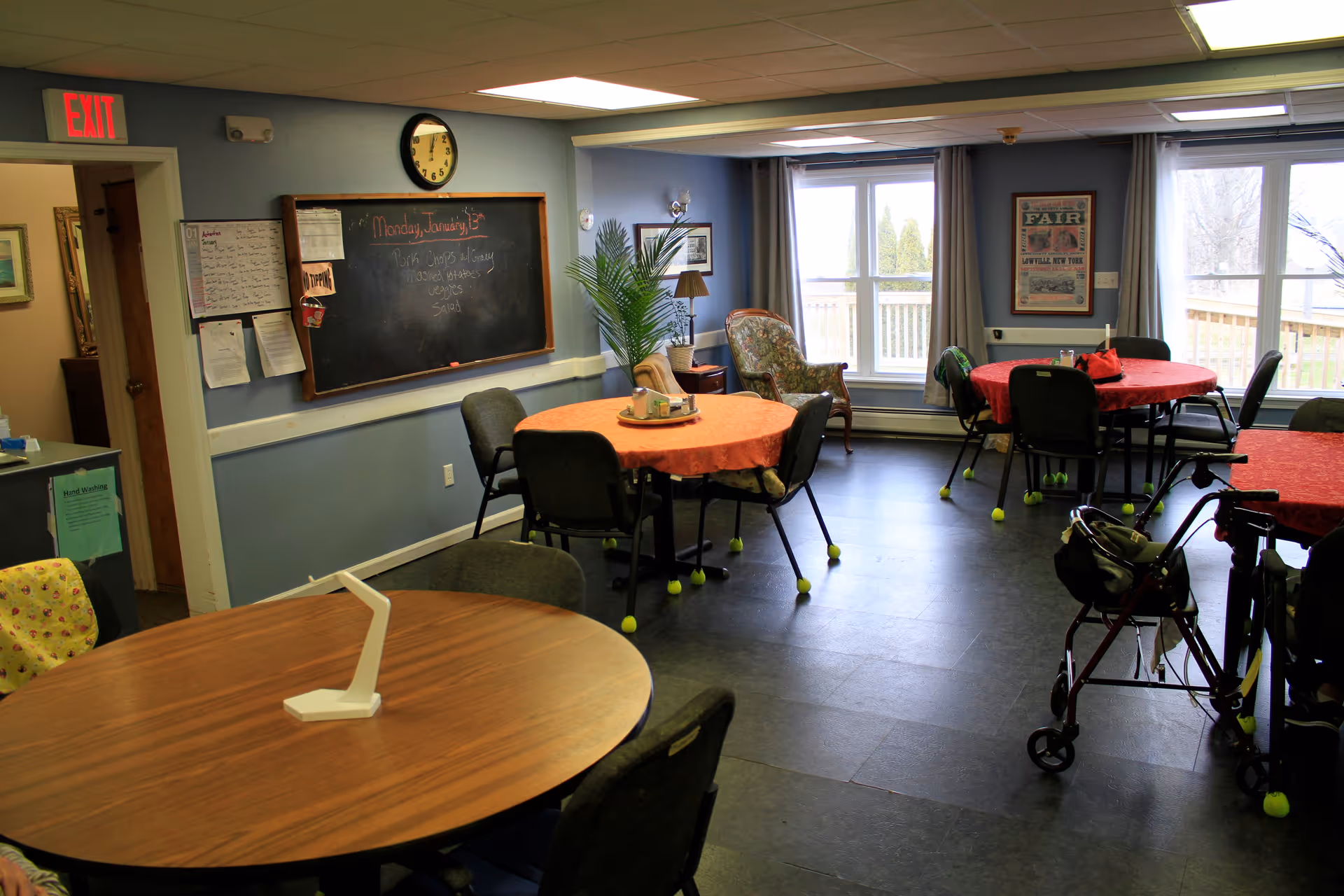 A common room in a senior living facility with several round tables covered with red and orange tablecloths, surrounded by chairs with tennis balls on the legs. There is a blackboard on the wall with writing, a clock above it, and a large window letting in natural light. A walker is visible near one of the tables, and there is a comfortable armchair near the window.