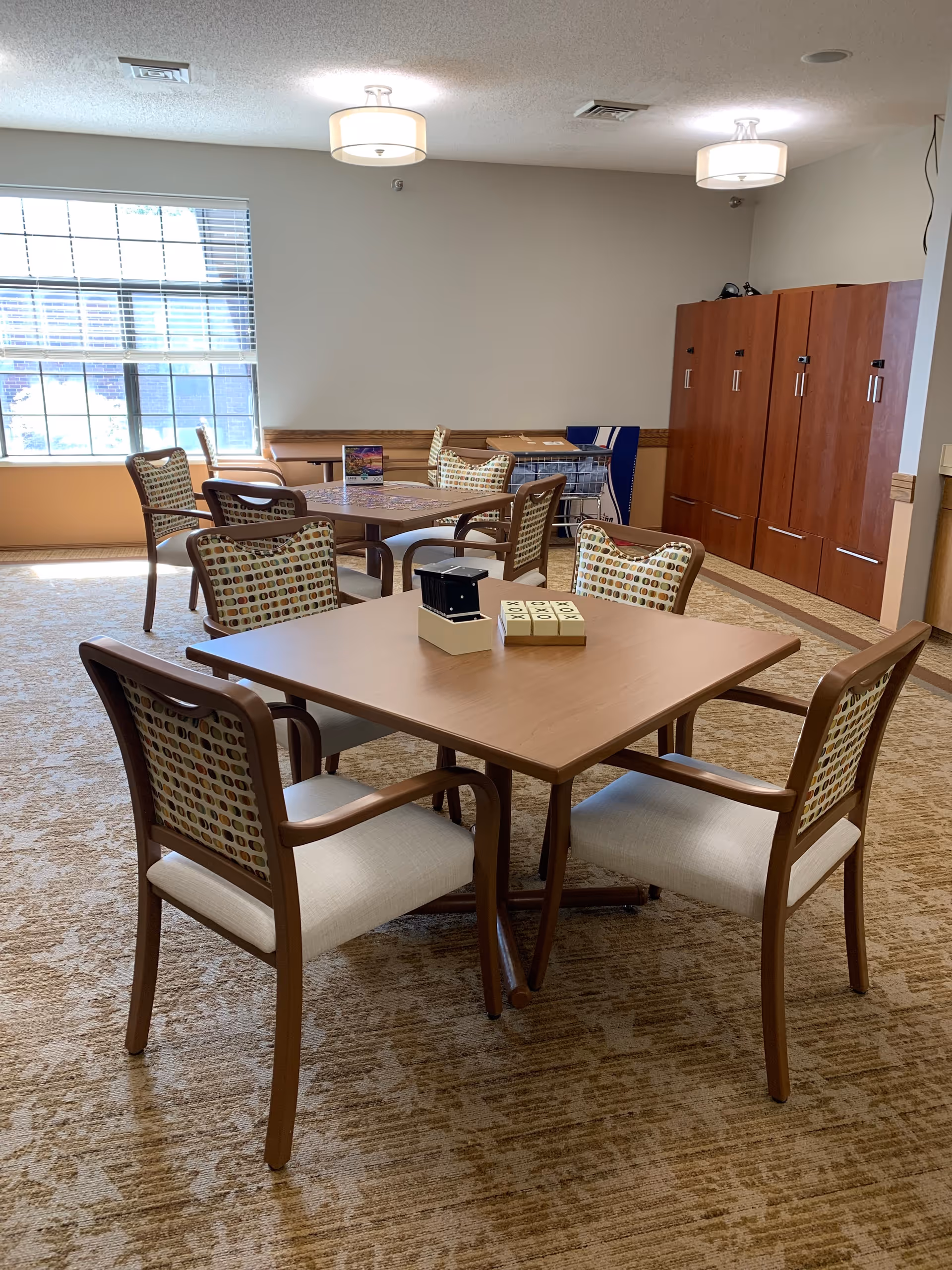 A well-lit room with multiple wooden tables and chairs arranged for seating. The chairs have patterned backs and light-colored cushions. There are two ceiling lights, a large window with blinds letting in natural light, and wooden cabinets along one wall. The carpet has a beige pattern.