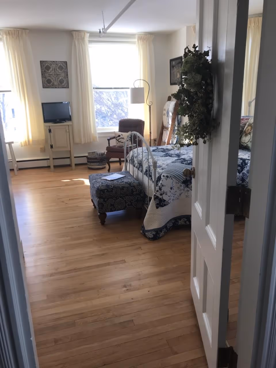 View into a bright bedroom with wooden floors, a bed with a blue and white quilt, a matching ottoman, a cushioned armchair, a floor lamp, a small TV on a stand, and two windows with cream-colored curtains. A wreath hangs on the partially open door.