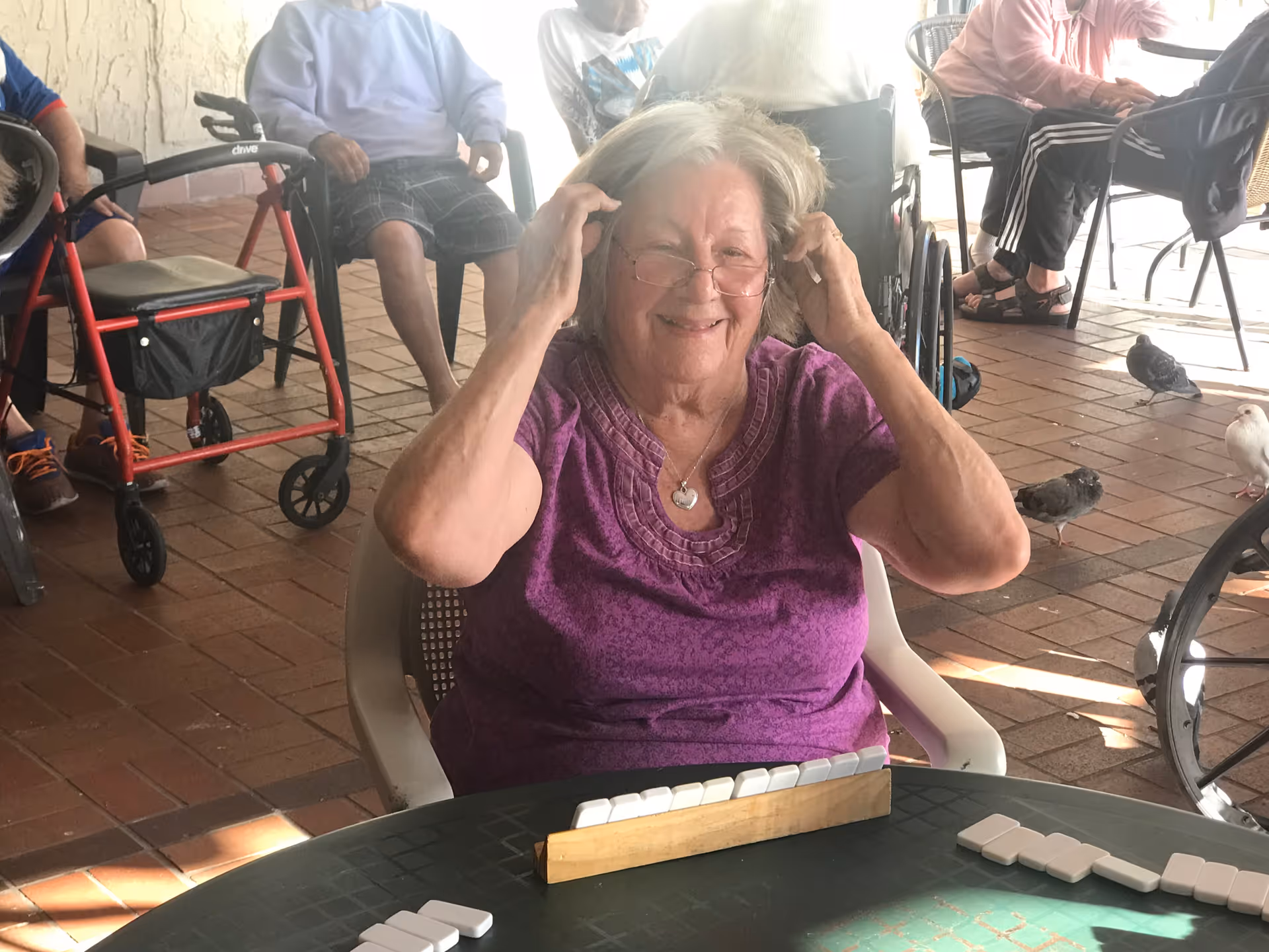 An elderly woman wearing glasses and a purple shirt is sitting at a table playing a game with white tiles, smiling and adjusting her glasses. Other elderly people are seated in the background, some in wheelchairs, on a brick-paved outdoor patio with birds nearby.