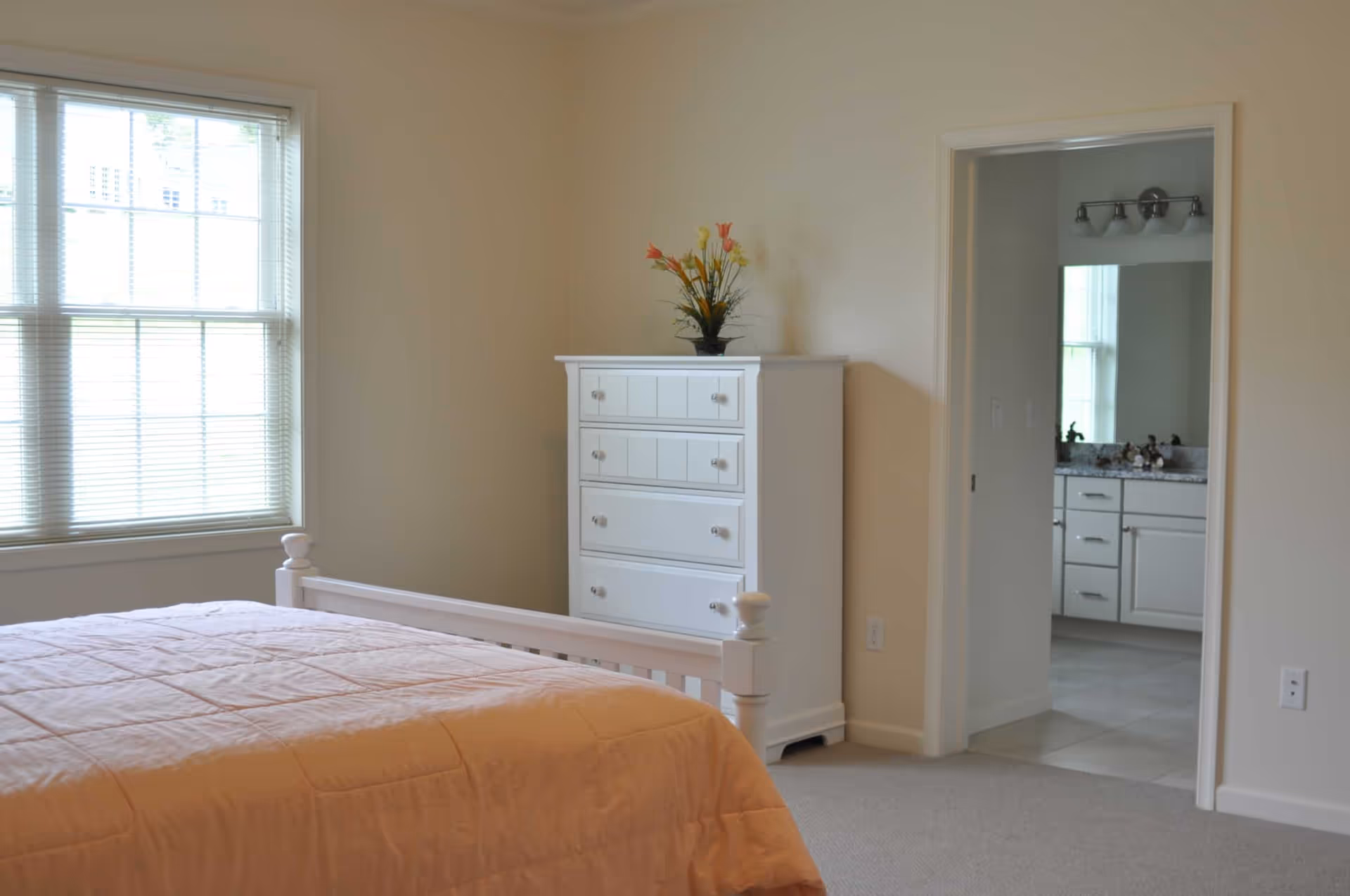 A bedroom with a large window with blinds on the left, a white wooden bed with a peach-colored quilt, and a white chest of drawers with a vase of flowers on top. An open doorway on the right leads to a bathroom with a vanity and mirror.