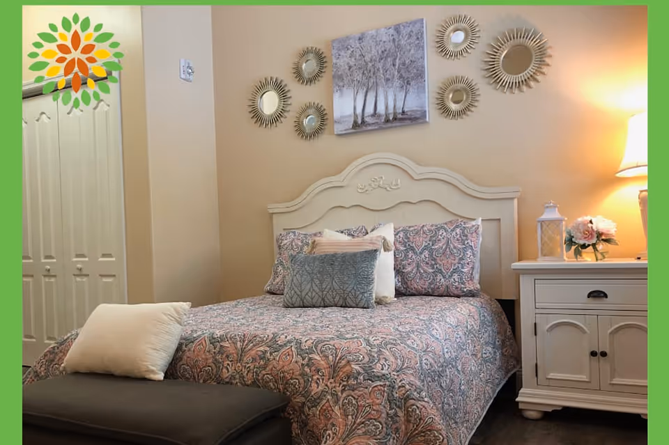 A cozy bedroom with a white wooden headboard bed adorned with patterned bedding and multiple pillows. Above the bed, there are decorative sunburst mirrors and a painting of trees on the wall. To the right of the bed is a white nightstand with a lamp, a lantern, and a vase with flowers. A closed closet with double doors is visible on the left side of the room.