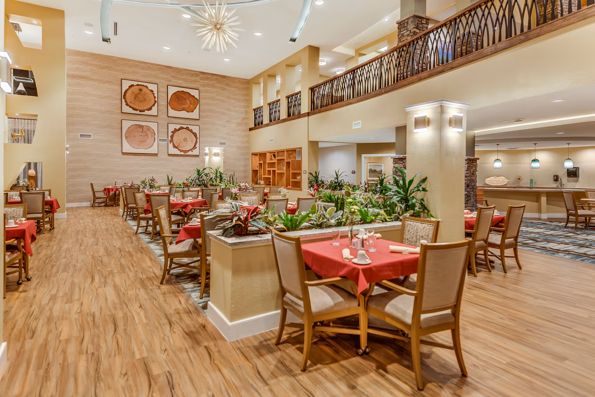 Spacious dining room with wooden flooring, tables covered with red tablecloths, and beige cushioned chairs. The room features a high ceiling with modern light fixtures, decorative wall art resembling wood slices, and a variety of green plants placed on dividers between seating areas. There is a balcony with wrought iron railing overlooking the dining area.