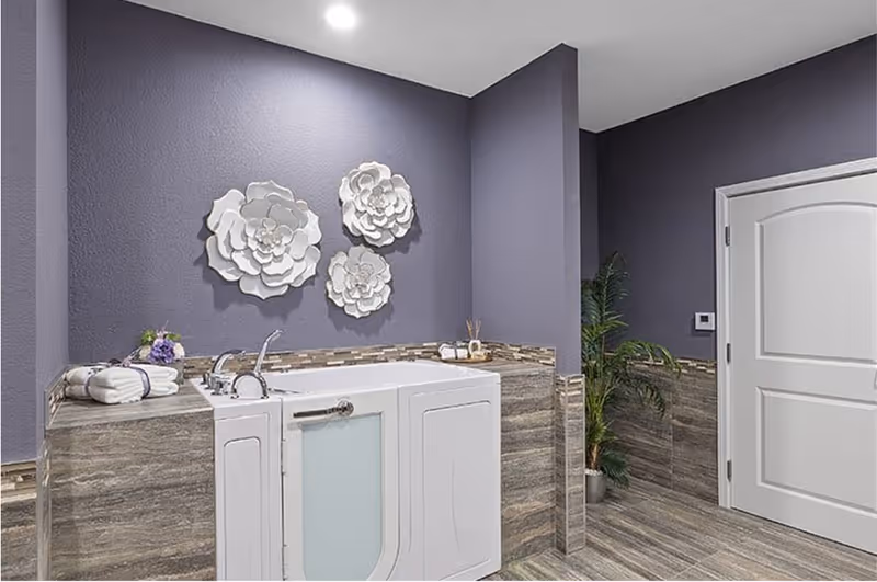 A walk-in bathtub with a white door and silver faucet set against a purple wall with decorative white flower wall art. The bathtub surround and floor have a wood-like tile finish. Towels and a small flower arrangement are placed on the tub ledge, and a potted plant is visible near a closed white door.