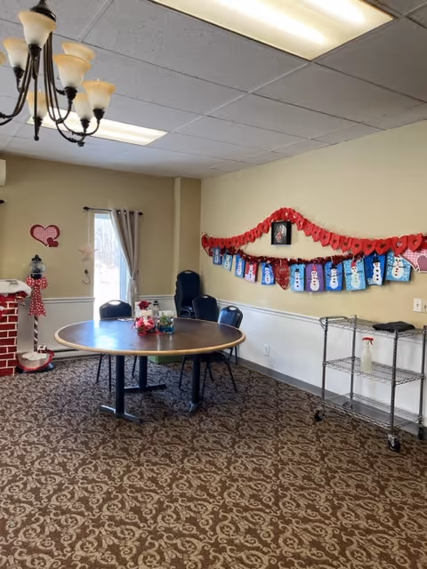 A cozy room with a round wooden table surrounded by black chairs on a patterned carpet. The walls are decorated with a red paper heart garland and blue and white snowman crafts. A window with beige curtains lets in natural light. There is a metal rolling cart with cleaning supplies on the right side and a decorative red brick chimney with a red and white polka dot scarf on the left.