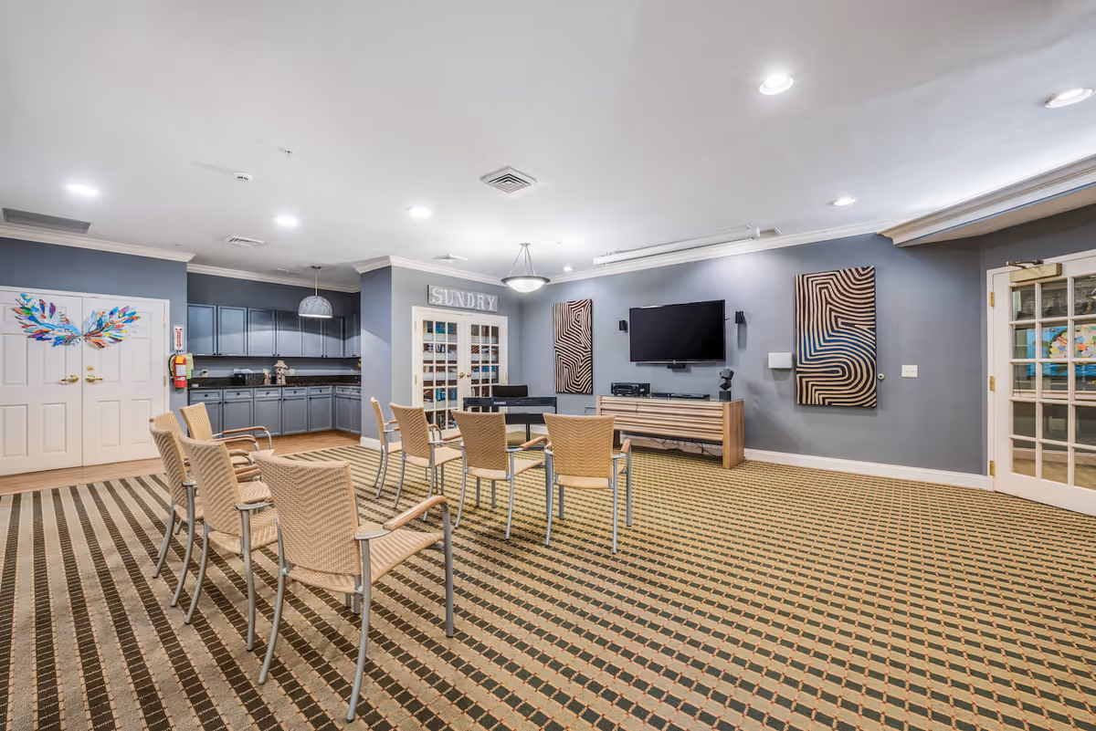 A spacious common room with a patterned carpet, several wicker chairs arranged in rows, a wall-mounted flat-screen TV, abstract wall art, and a kitchenette area with gray cabinets. There are double doors with colorful wing artwork on the left and a glass-paneled door on the right. A sign above a set of glass doors reads 'SUNDRY'.
