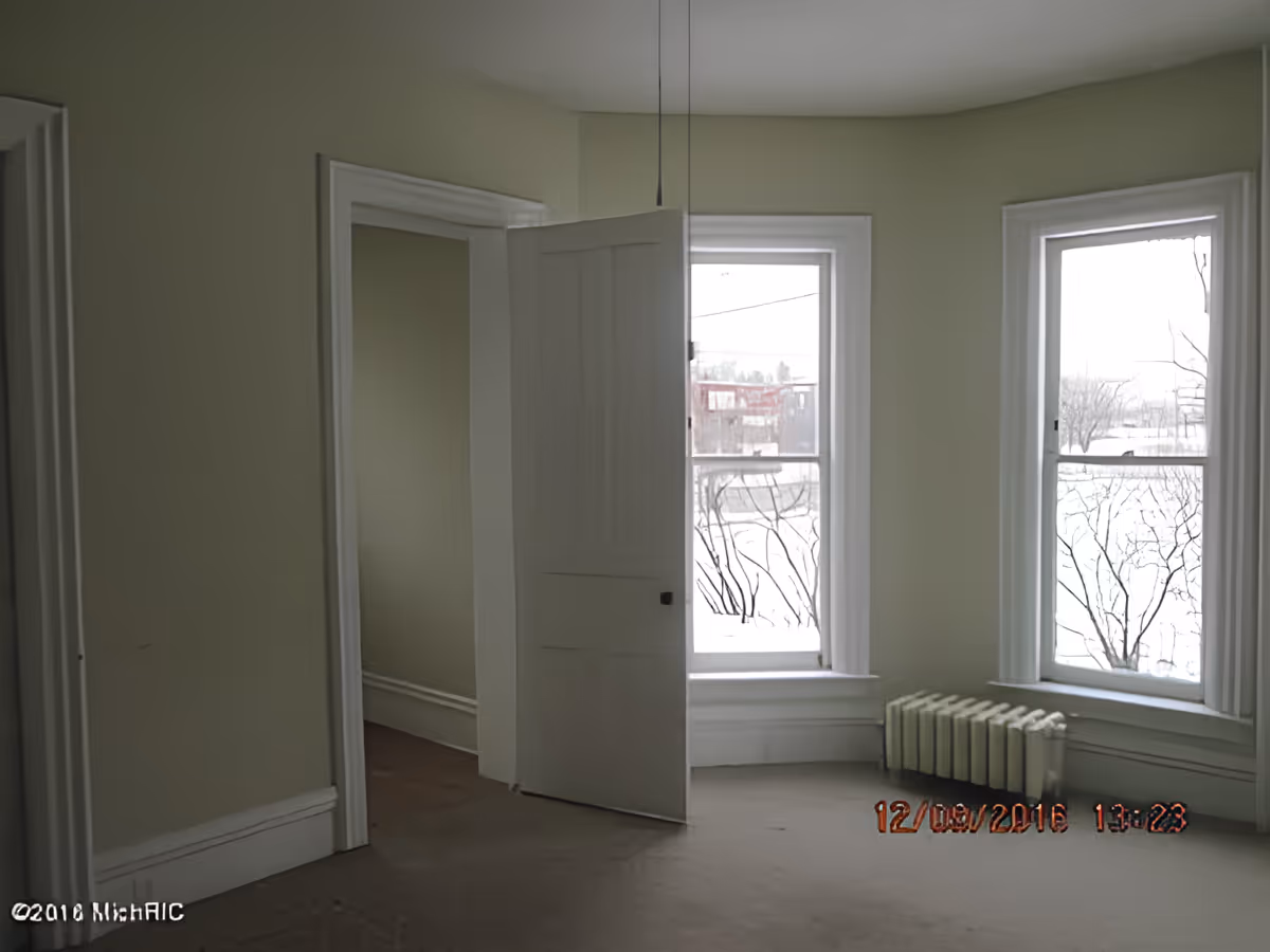 Empty carpeted room with two tall windows, a radiator beneath one window, and an open interior door.
