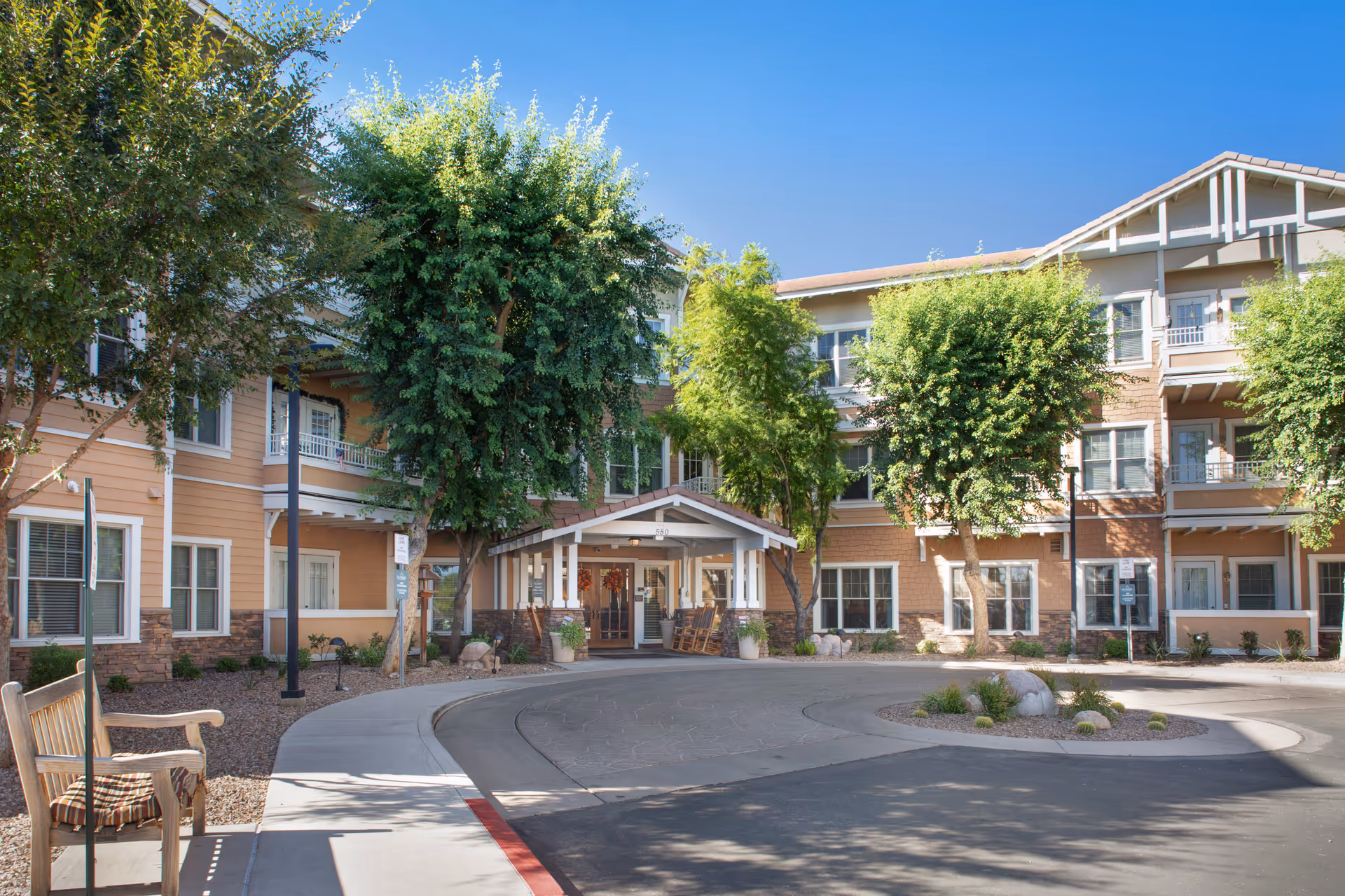Exterior view of Sunrise of Gilbert senior living facility with a circular driveway, trees, and a covered entrance with rocking chairs. The building is three stories tall with beige and brown siding and white trim.