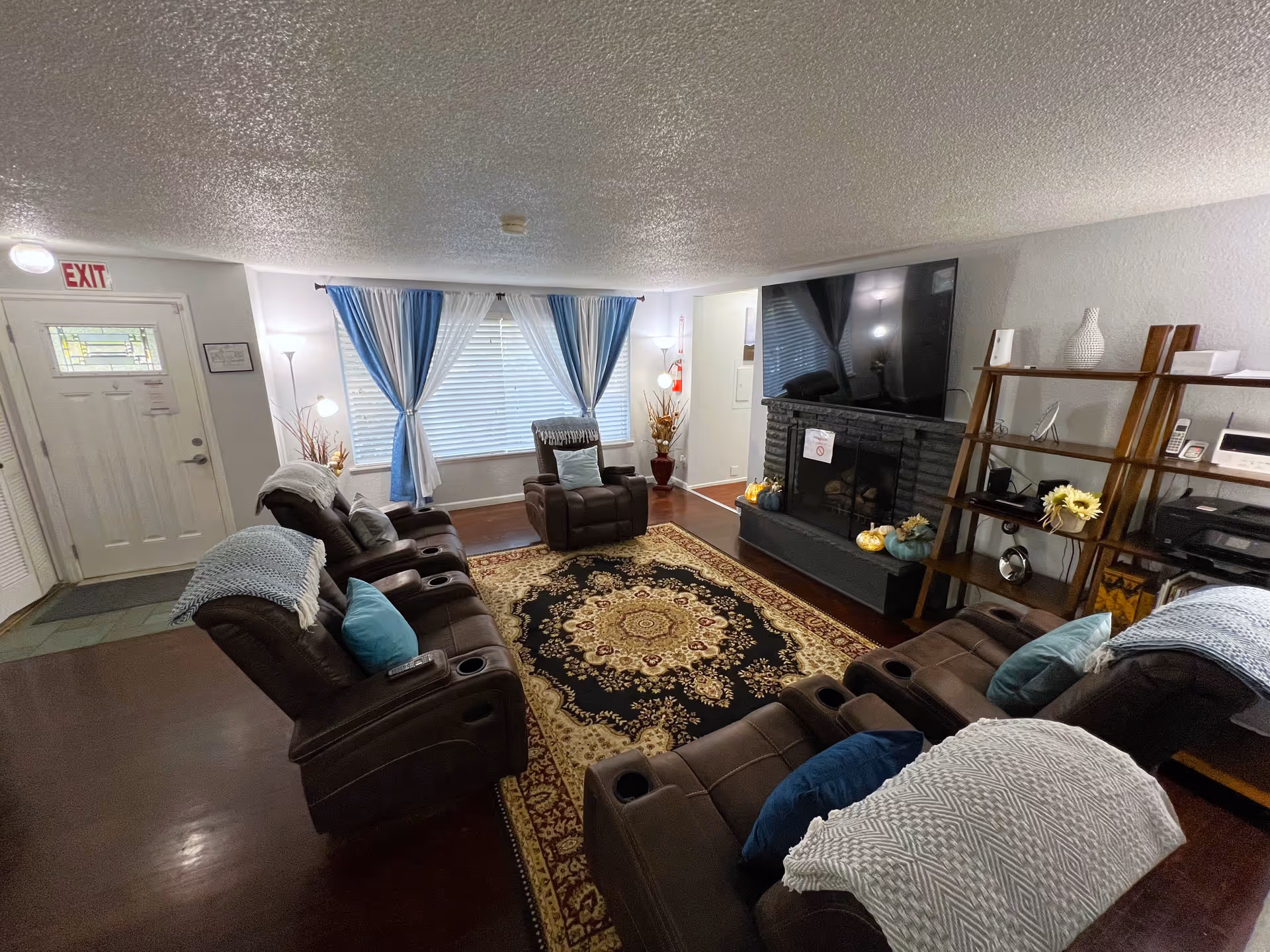 A cozy living room with brown reclining chairs arranged around a patterned area rug. There is a large flat-screen TV mounted above a black fireplace decorated with small pumpkins and flowers. Blue and white curtains frame a large window, and a wooden shelving unit with various items is visible on the right. The entrance door with an exit sign above it is on the left side of the room.