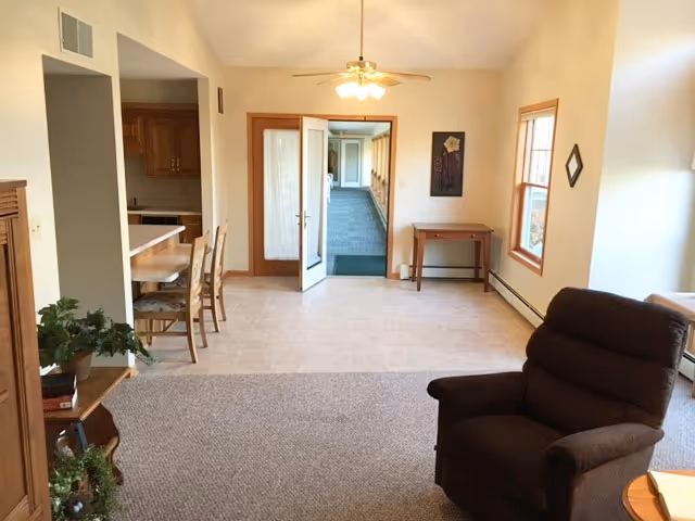 Interior view of a senior living facility room with a brown recliner chair in the foreground, a small wooden table with a plant on the left, a dining area with wooden chairs and a counter on the left side, and a door leading to a hallway in the background. The room has beige walls, carpet and tile flooring, and windows letting in natural light.