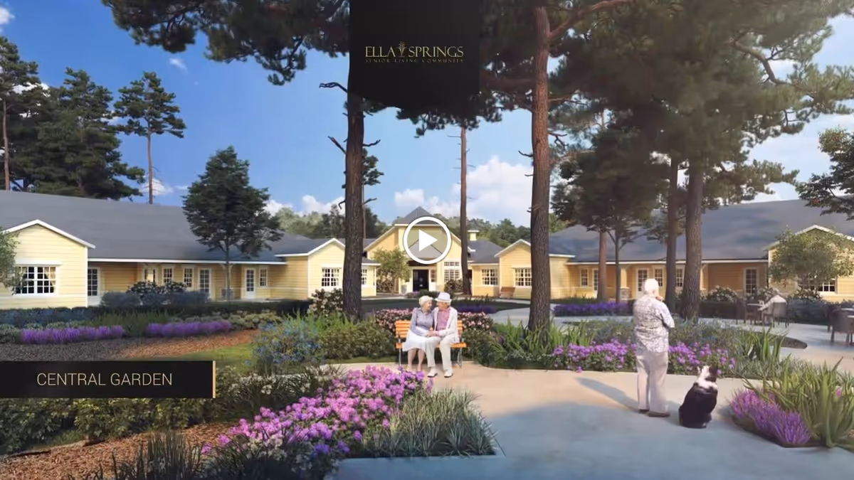 A peaceful central garden area at Ella Springs Senior Living Community featuring well-maintained flower beds with purple and green plants, tall pine trees, and yellow single-story buildings in the background. Two elderly women are sitting on a bench having a conversation, while another elderly person stands nearby with a black and white dog. The sky is clear and blue, creating a serene outdoor environment.