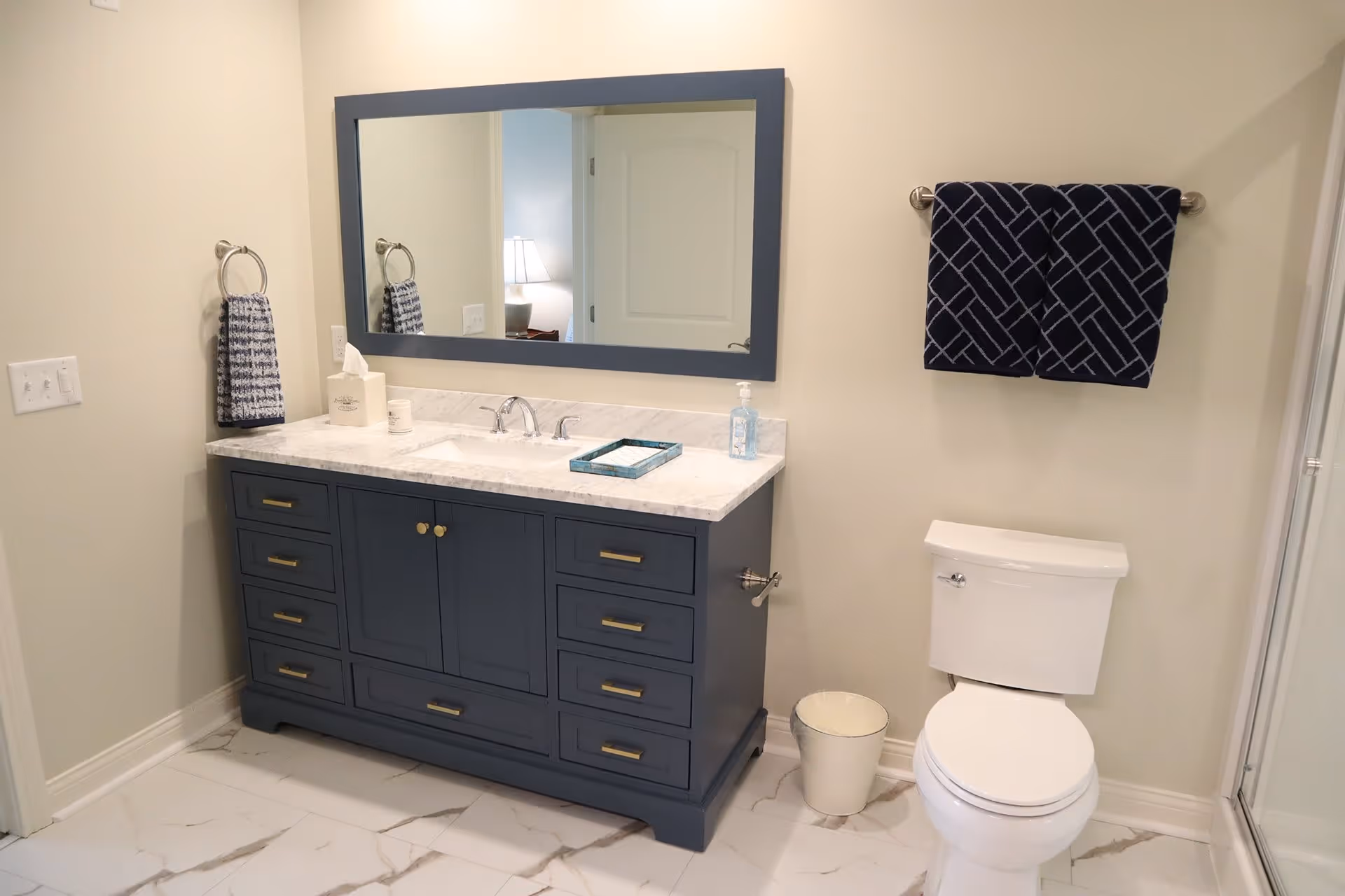A clean and modern bathroom featuring a blue vanity with a white marble countertop, a large rectangular mirror above it, a white toilet, and two dark blue patterned towels hanging on silver towel racks. The floor is tiled with white marble tiles with gray veining.