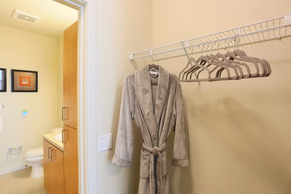 A beige bathrobe hanging on a wire closet rack with several empty hangers. To the left, there is an open door leading to a bathroom with a toilet, sink, wooden cabinets, and two framed pictures on the wall.