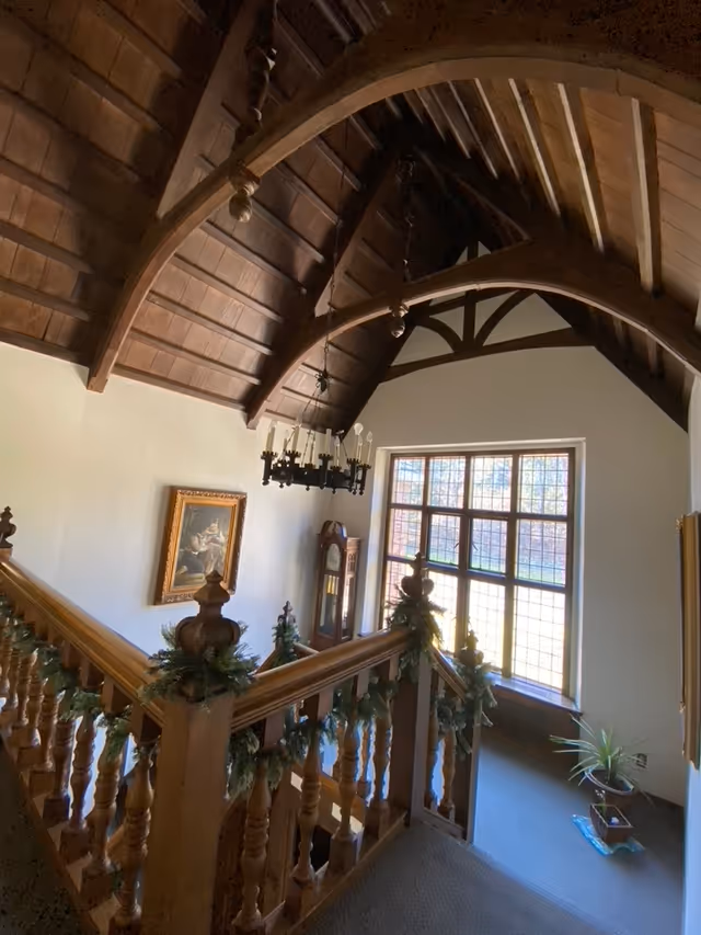 Interior view of a stairway area with wooden railings decorated with greenery. The ceiling features dark wooden beams and a chandelier. A large window with multiple panes lets in natural light. There is a grandfather clock and a framed painting on the wall, and a potted plant near the window.