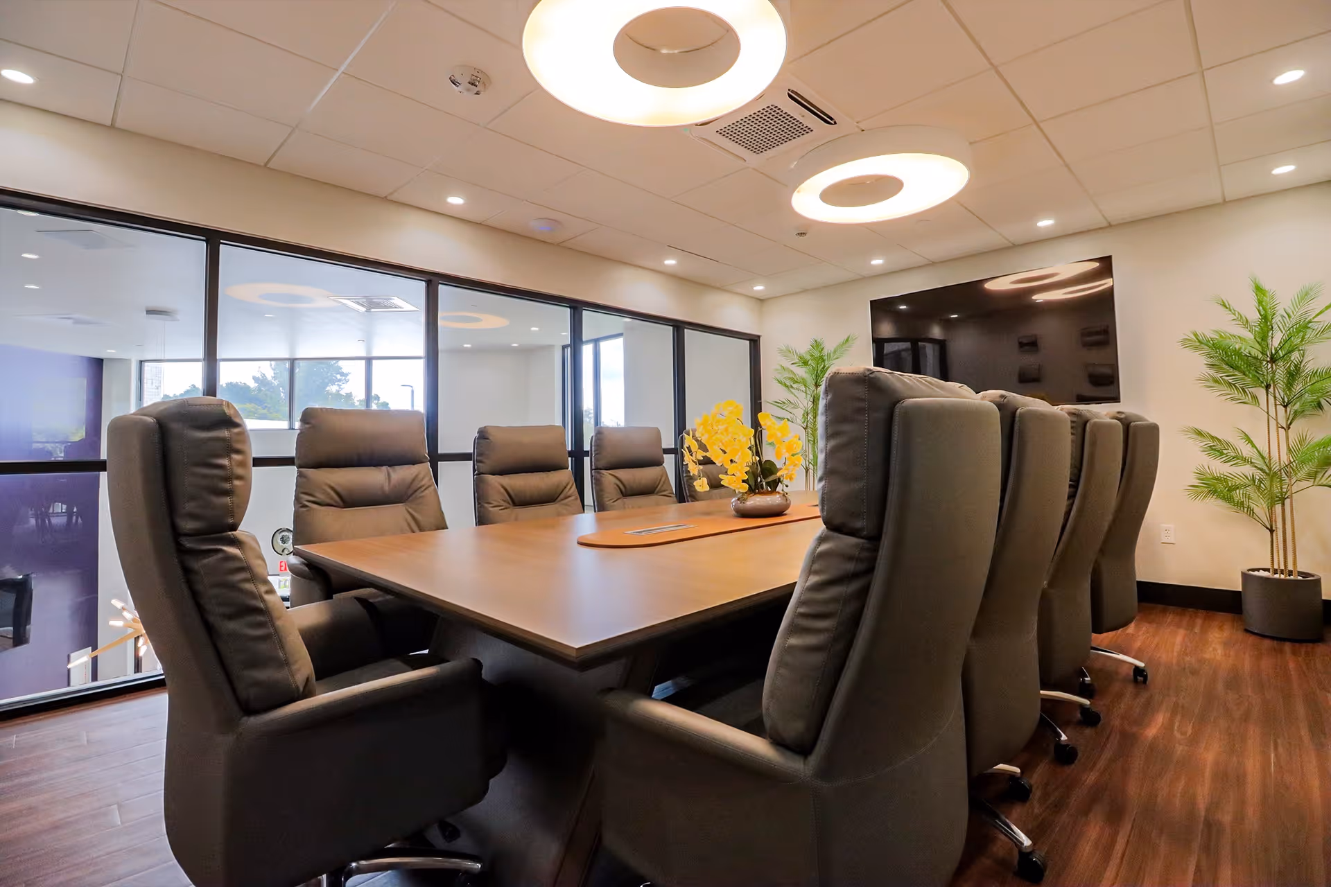 Conference room with a long wooden table surrounded by high-backed leather chairs, a wall-mounted TV, and potted plants.