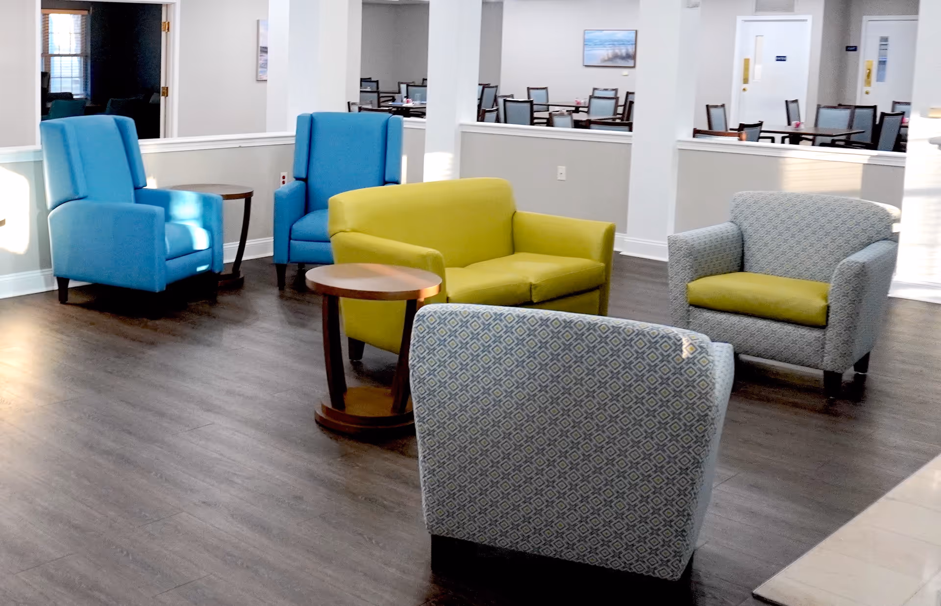 A seating area in a senior living facility with two blue armchairs, a green loveseat, and two patterned armchairs with green cushions arranged around two small wooden side tables. In the background, there is a dining area with tables and chairs, and light-colored walls with framed artwork.