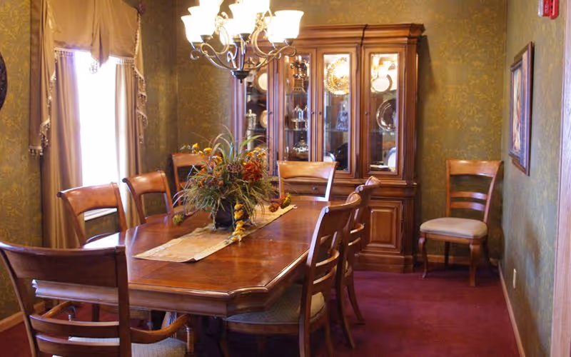 Traditional dining room with a wooden table and chairs, floral centerpiece, chandelier, and a glass-front china cabinet.