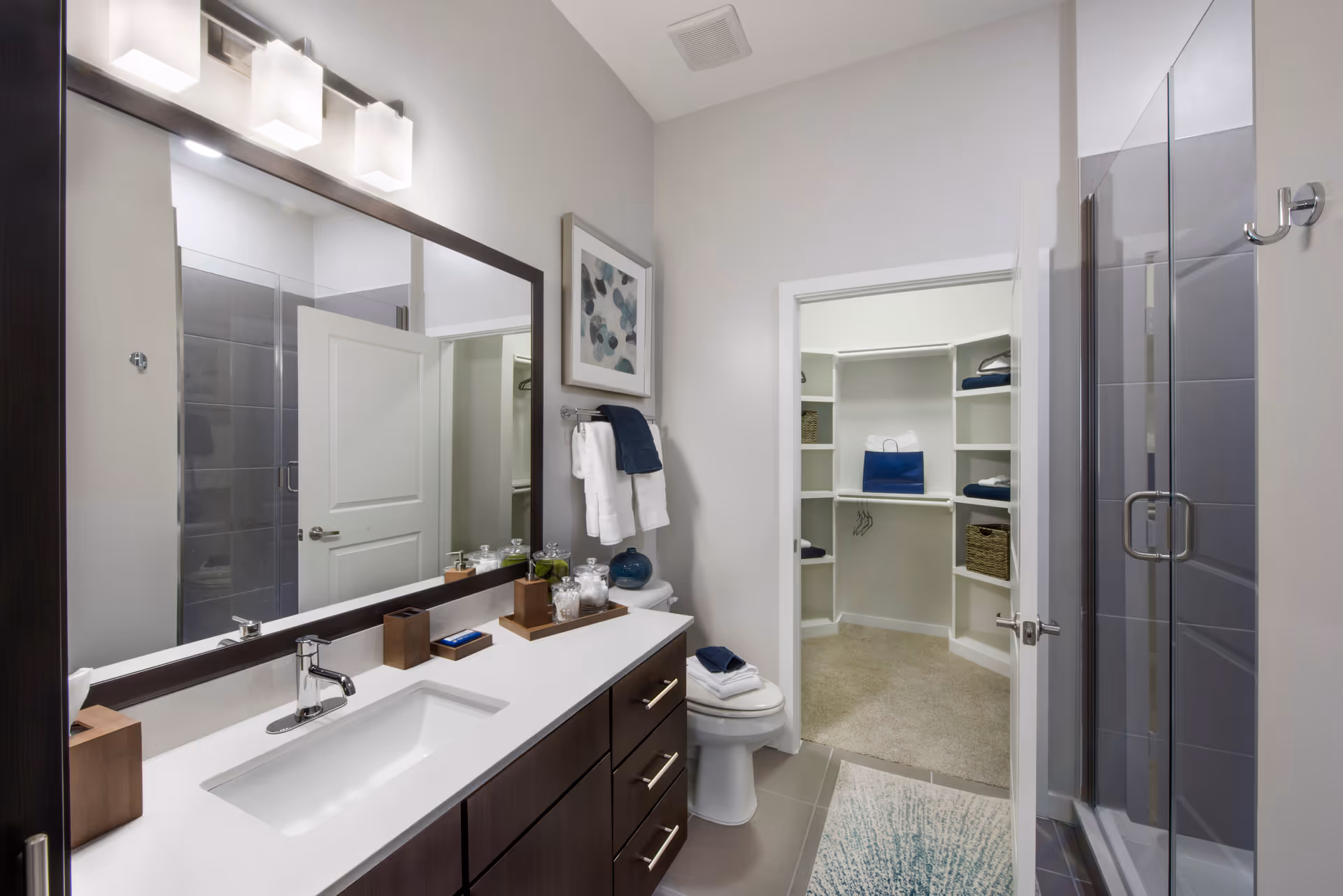 Modern bathroom with a large mirror above a white countertop and dark wood cabinets. The bathroom features a sink, a toilet with folded towels on top, a glass-enclosed shower with gray tiles, and a door leading to a walk-in closet with shelves and hanging space. The walls are light-colored, and there is a framed abstract artwork above the towel rack.