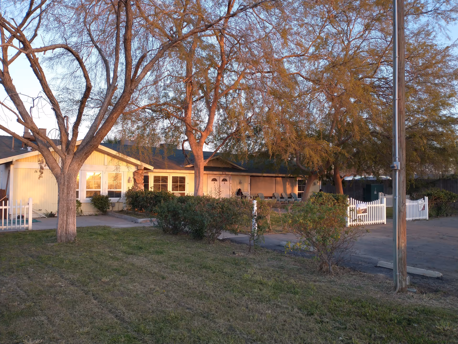Exterior view of a single-story building with a green roof and light yellow walls, partially obscured by leafless trees and bushes. There is a white picket fence and a paved driveway or parking area in front. The scene is lit by warm sunlight, suggesting early morning or late afternoon.