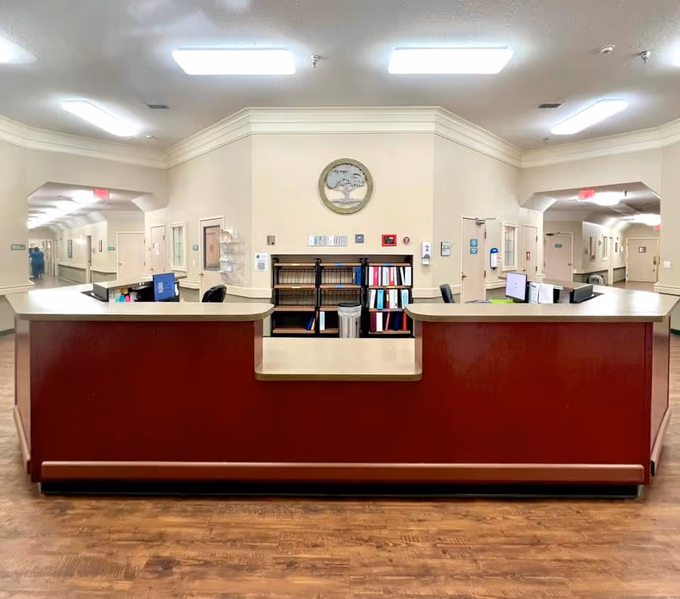Reception desk area inside a nursing and rehabilitation center with a maroon front counter, computer monitors, office chairs, shelves with binders, and a decorative wall clock above. Hallways extend on both sides with doors and lighting fixtures on the ceiling.