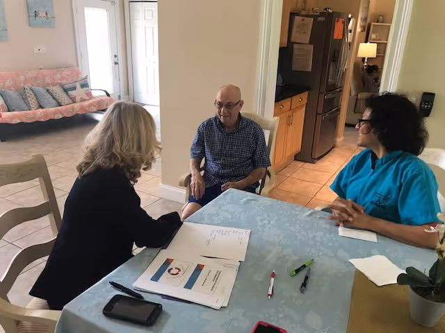 Three people sitting around a table in a residential-style room. One elderly man is seated in the middle, with a woman in a blue uniform on the right and another woman with blonde hair on the left. The table has papers, pens, and a smartphone on it. In the background, there is a couch with cushions and a door leading outside.