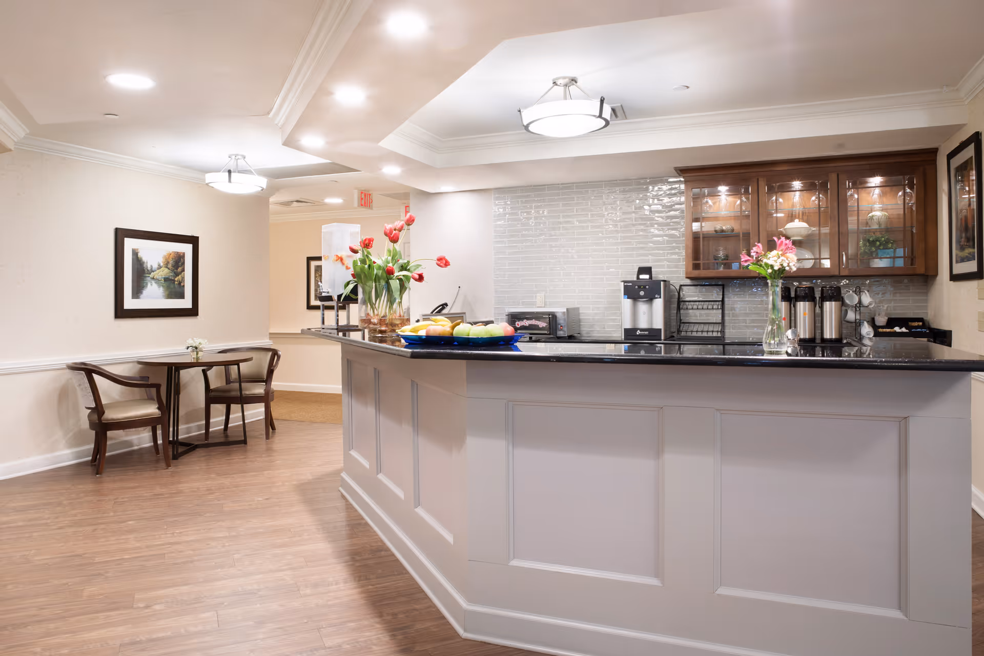 A bright and clean common area with a large counter featuring a black countertop, a coffee machine, and vases with flowers. Behind the counter are wooden cabinets with glass doors displaying decorative items. To the left, there is a small seating area with a round table and two chairs, and framed artwork on the wall. The floor is wooden, and the ceiling has recessed lighting and a modern light fixture.