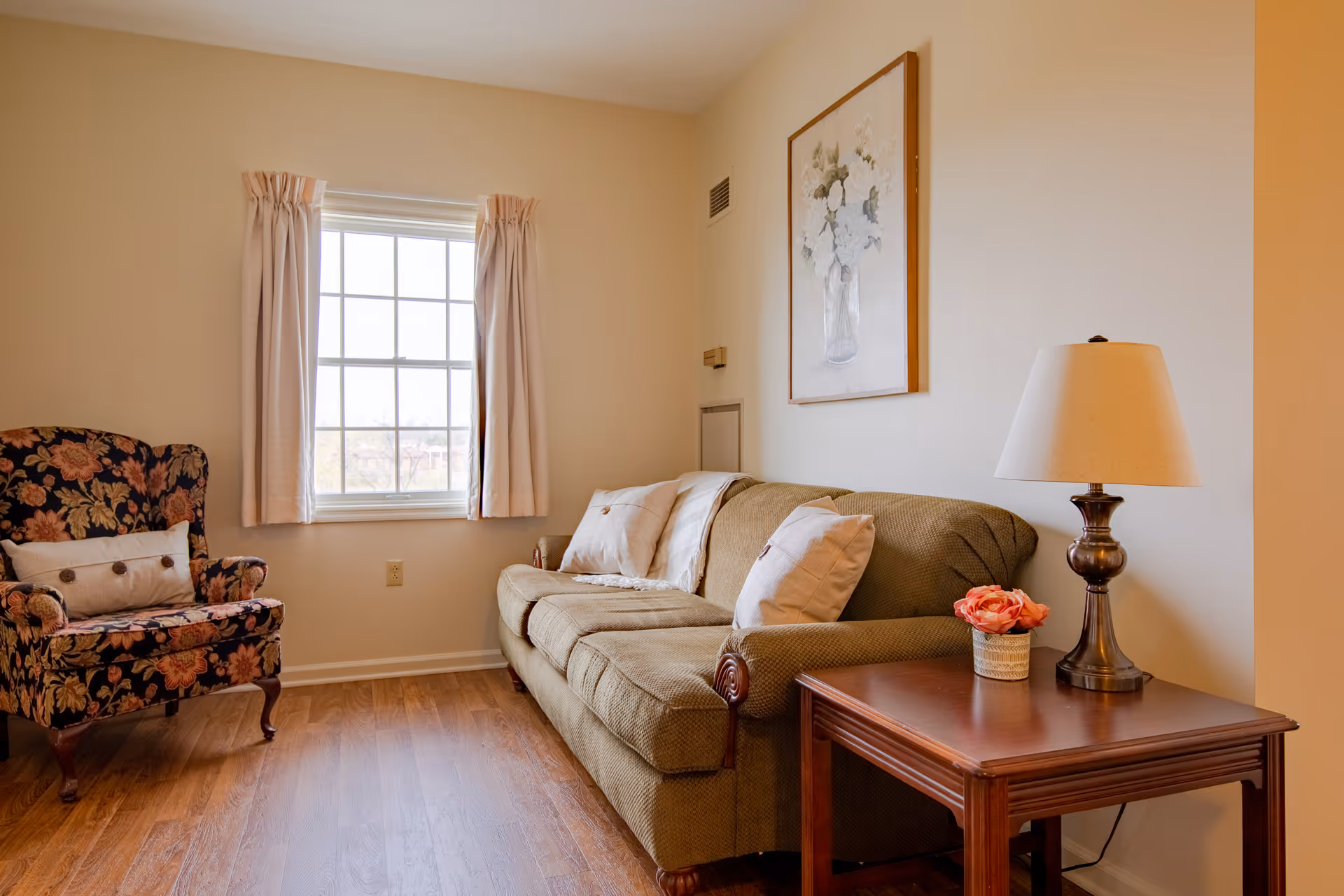 A cozy living room with a green sofa, floral armchair, side table with a lamp and flower, and a window with curtains.
